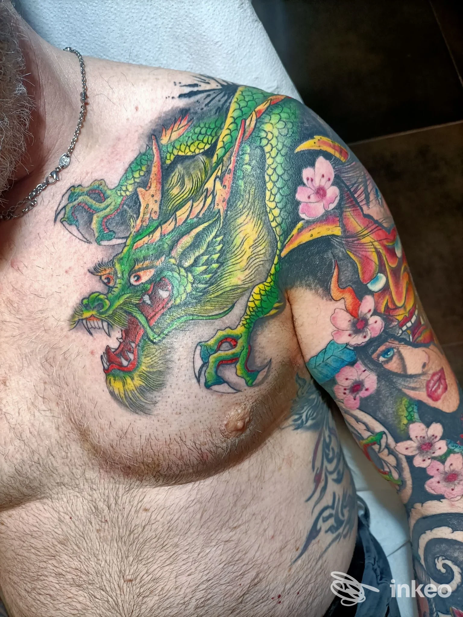 Tatuaggio colorato con grande drago verde su petto, spalla e braccio e fiori rosa; lavoro di Gigi Tattoo a Montecchio Maggiore.