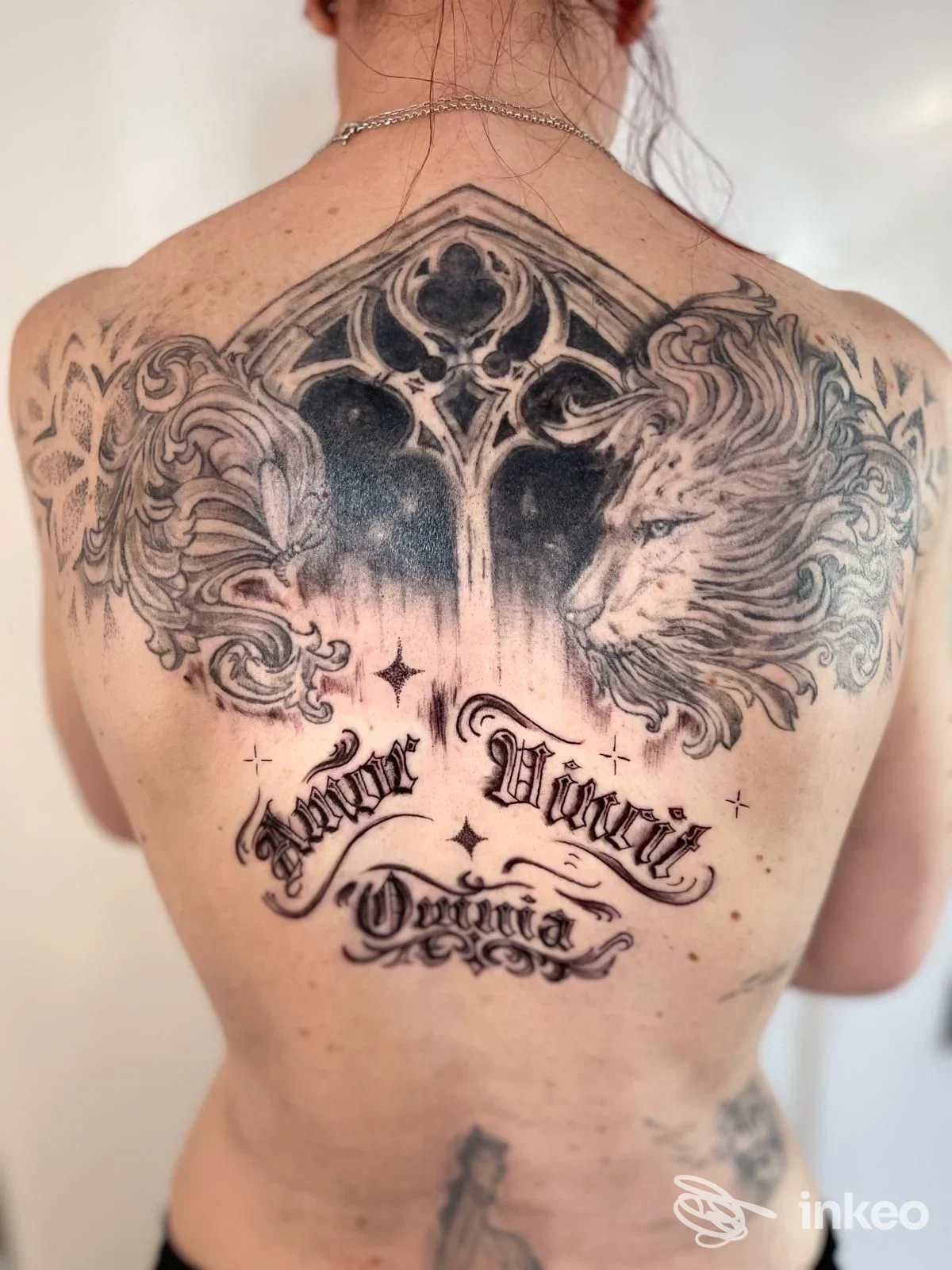 Grande tatuaggio in bianco e nero sulla schiena con architettura ornamentale, testa di leone e scritte; immagine fornita da INK ME Tattoo, Caldogno.