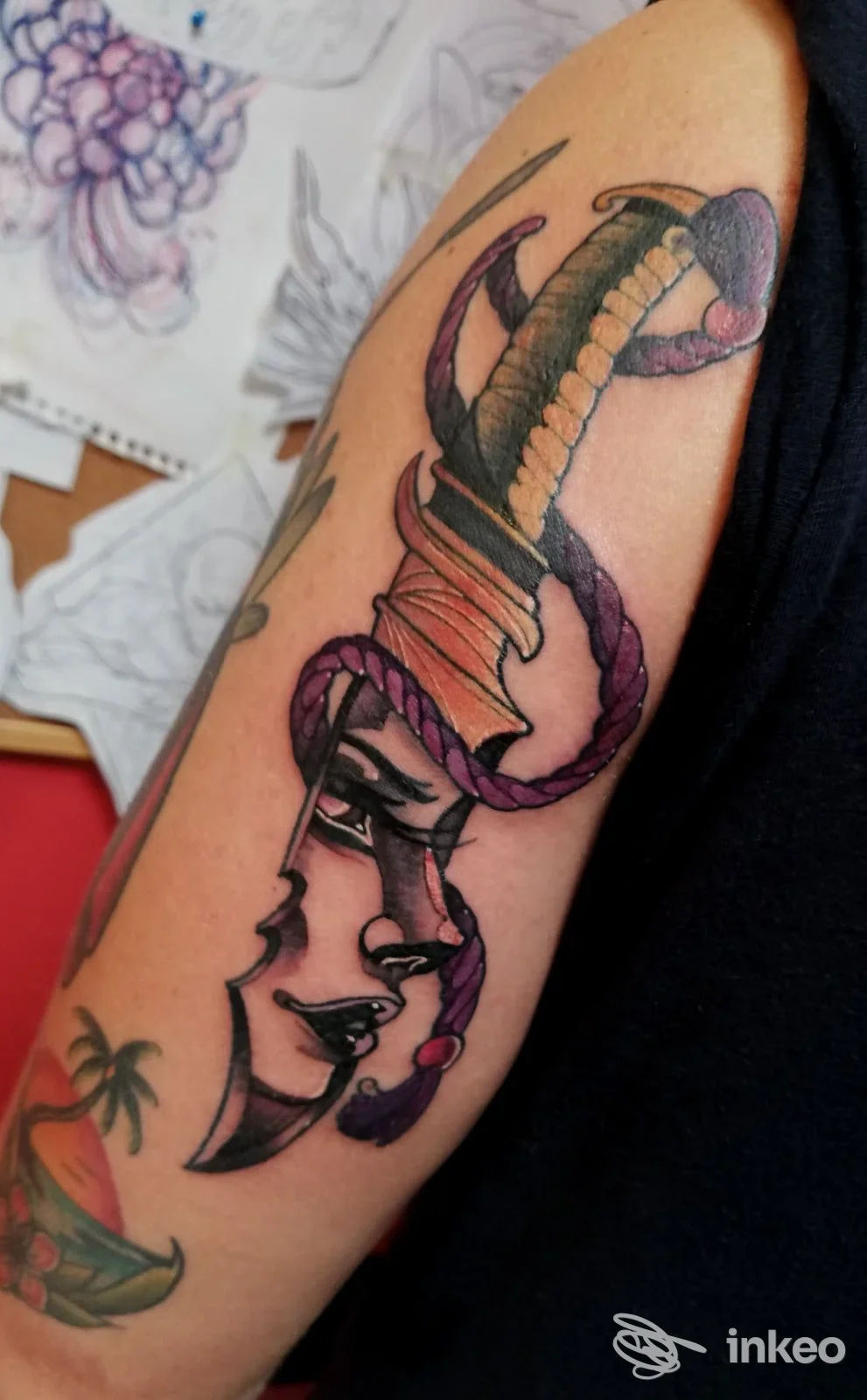Tatuaggio a colori su braccio superiore con pugnale e volto stilizzato, IV DIMENSIONE Tattoo Studio, Treviso.