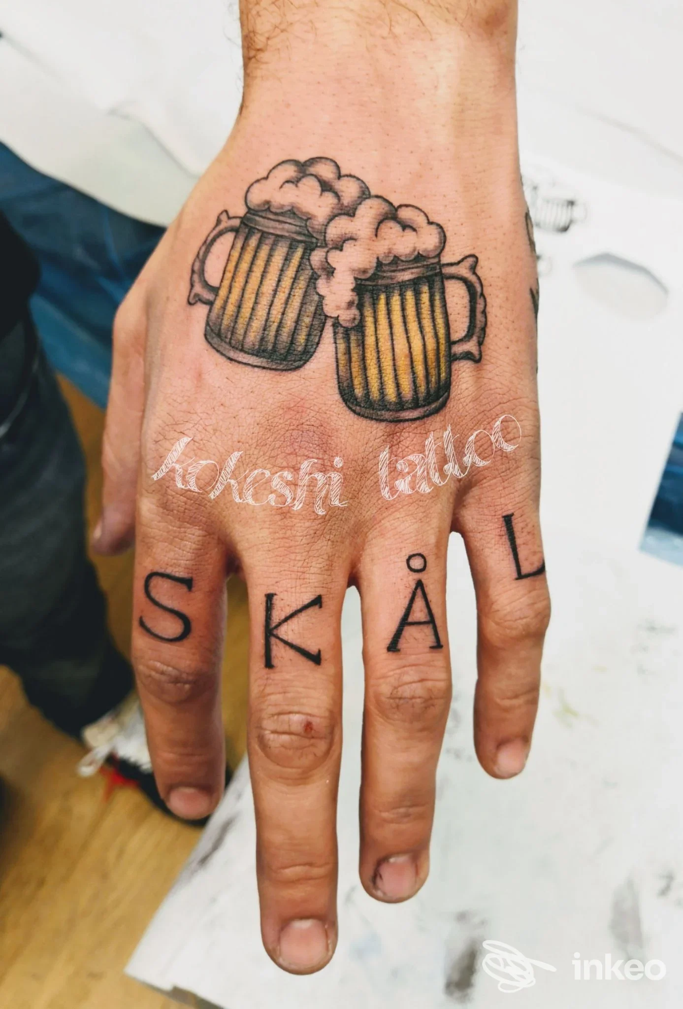 Mano tatuata con due boccali di birra sul dorso e scritte 'SKÅL' sulle dita. Studio KOKESHI TATTOO, San Zenone degli Ezzelini.