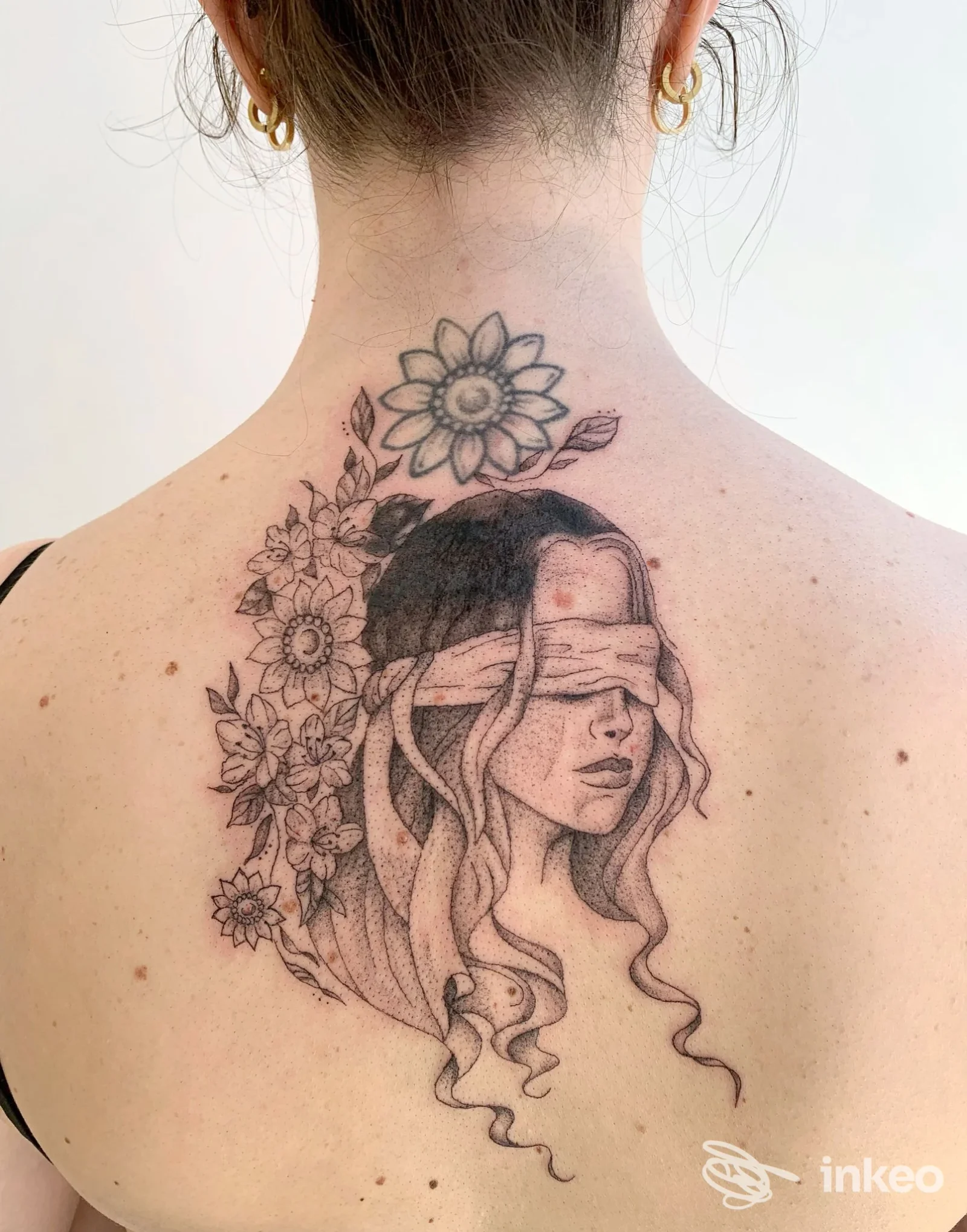 Grande tatuaggio in bianco e nero sulla nuca e parte superiore della schiena: ritratto femminile bendato con fiori. Libre Tattoo, Schio.