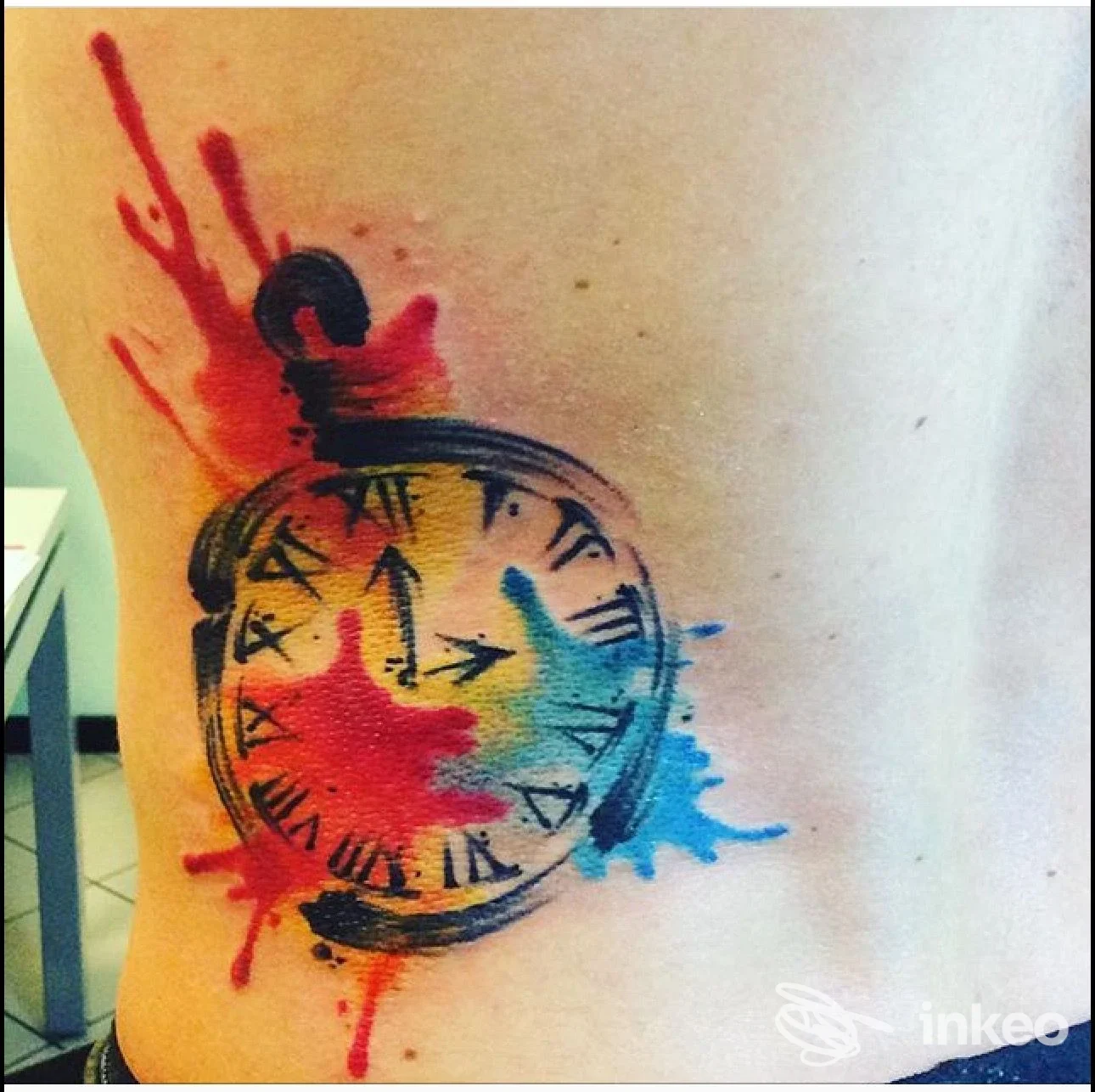 Foto di un tatuaggio acquerello a forma di orologio con schizzi rossi e blu sul fianco, realizzato da La Fenice Tattoo & Piercing, Belluno.