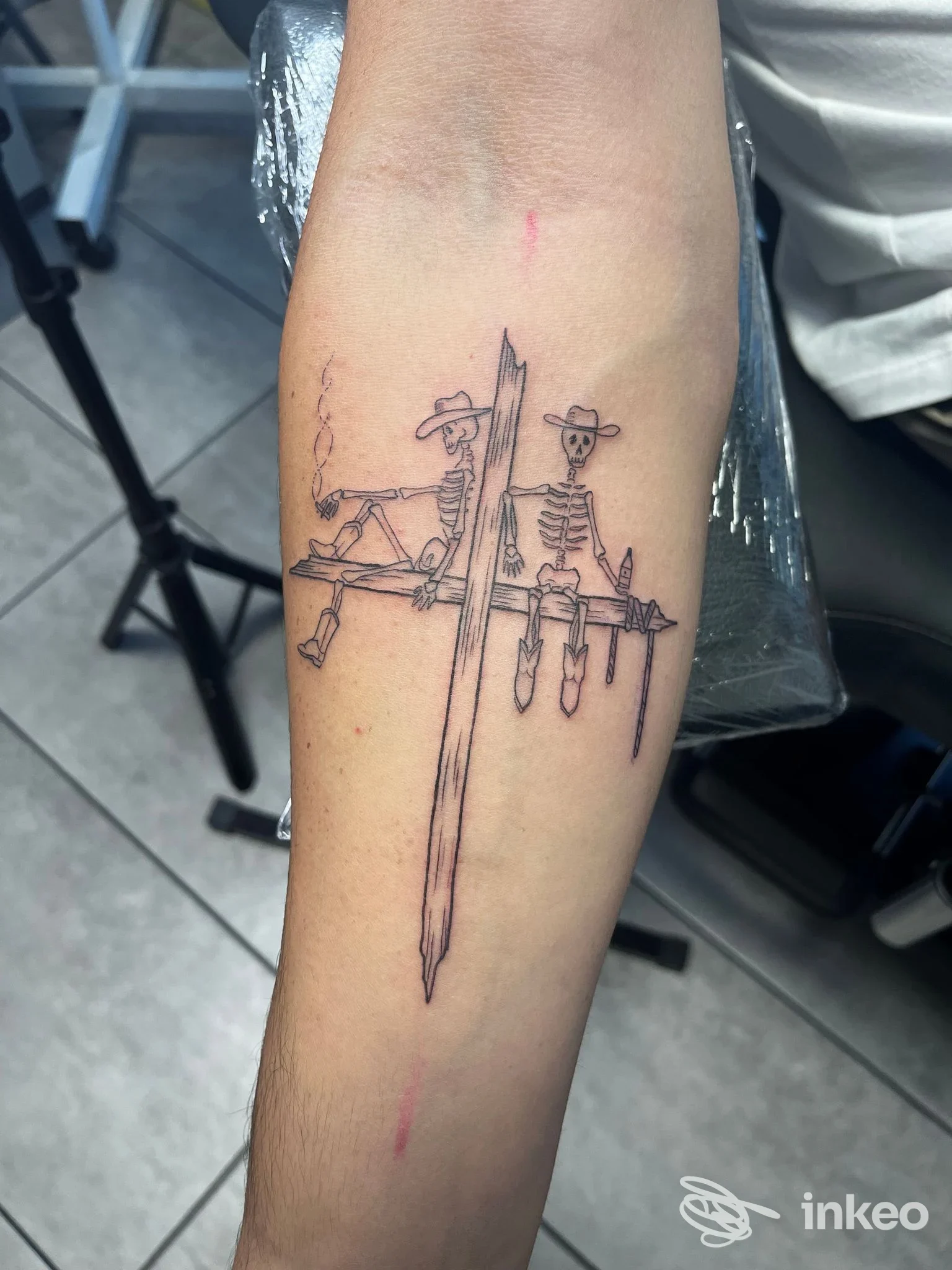Tatuaggio lineare su avambraccio con due scheletri e una spada centrale, realizzato presso Loose Doggs Tattoo Studio, Venezia.