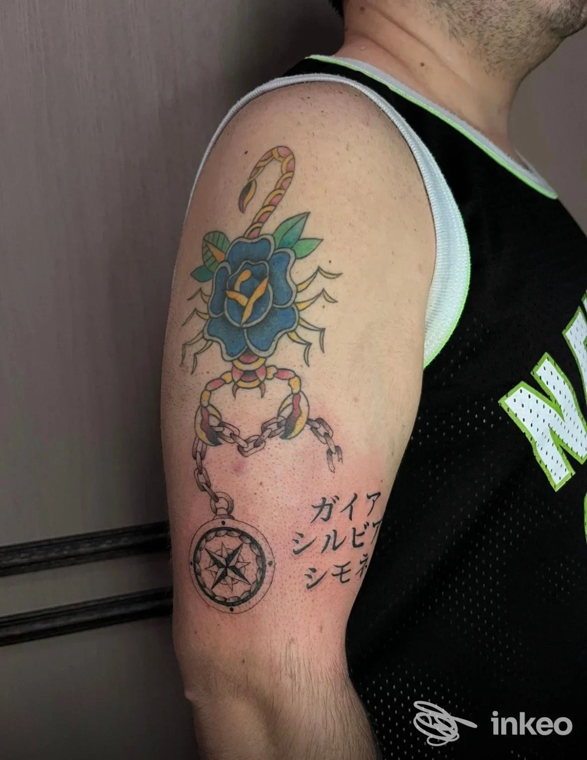 Tatuaggio colorato su braccio superiore con fiore blu, catena e ciondolo; foto inviata da Queen Of Ink Clinique Tattoo, Villa Fontana.
