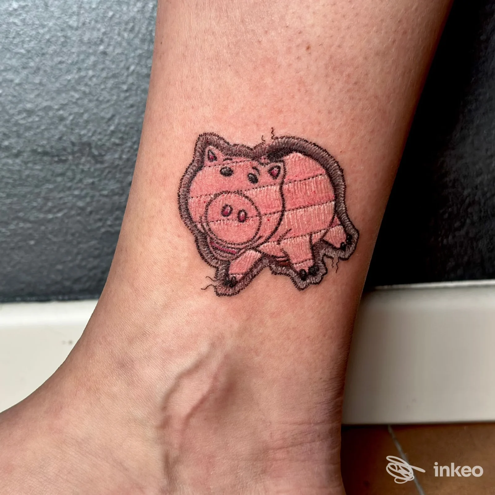 Tatuaggio di un piccolo maialino rosa con contorno scuro sulla caviglia, fotografato a Verona per Restart Tattoo.