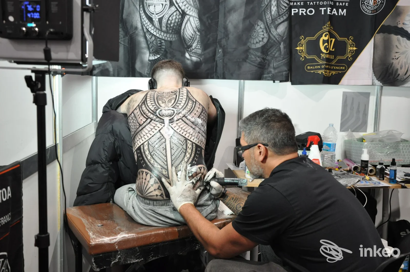 Scena in una convention di tatuaggi: una persona è seduta mentre un artista tattooing esegue un elaborato tatuaggio sul dorso, con strumenti e materiale sul tavolo.