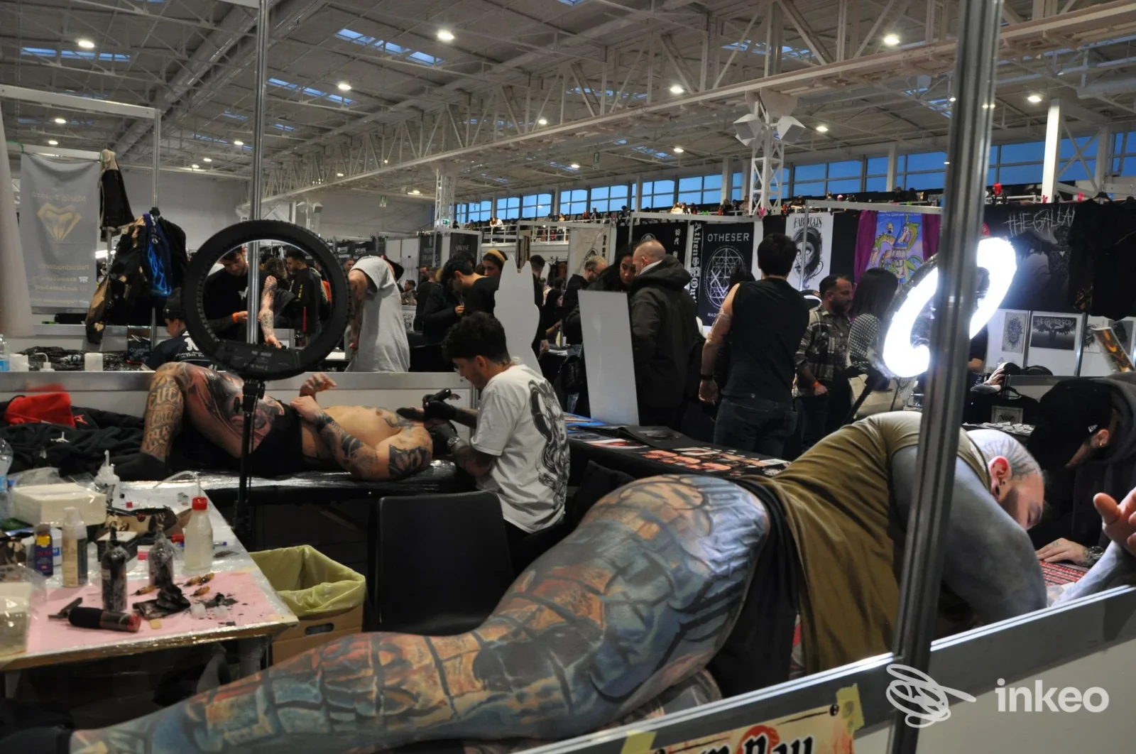 Milano Tattoo Convention: una persona è sdraiata mentre un’artista lavora su un grande tattoo su braccio e gamba, con pubblico e stand sullo sfondo in un padiglione luminoso.