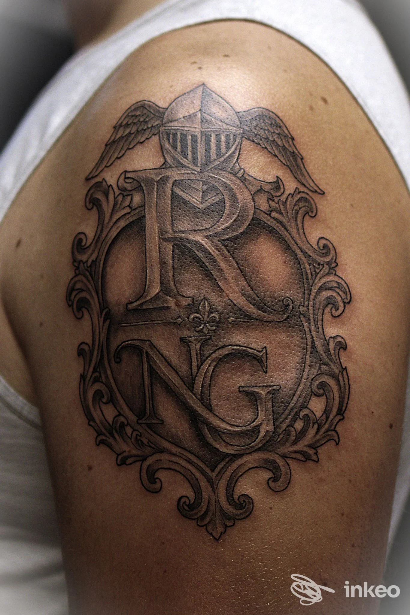 Grande tatuaggio black & grey sul deltoide con stemma ornamentale e lettere 'R' 'NG', immagine fornita da TATTOO STUDIO di Juri Scalco, Longare.