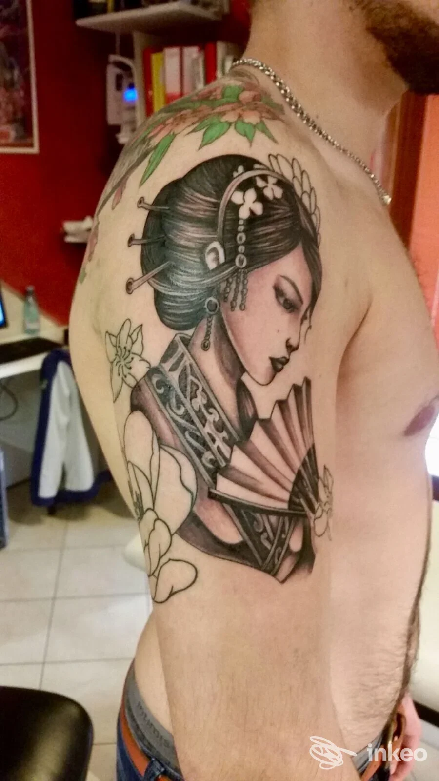 Tatuaggio sulla spalla di una geisha in profilo con ventaglio e fiori, fotografato su pelle di cliente. Armonia Tattoo, San Zeno.