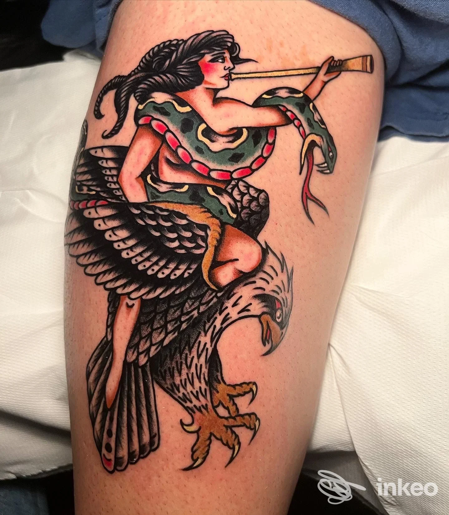 Tattoo colorato su coscia raffigurante una donna avvolta da un serpente e ali di un grande uccello, con tonalità rosse, nere e verdi, veduta frontale della gamba.