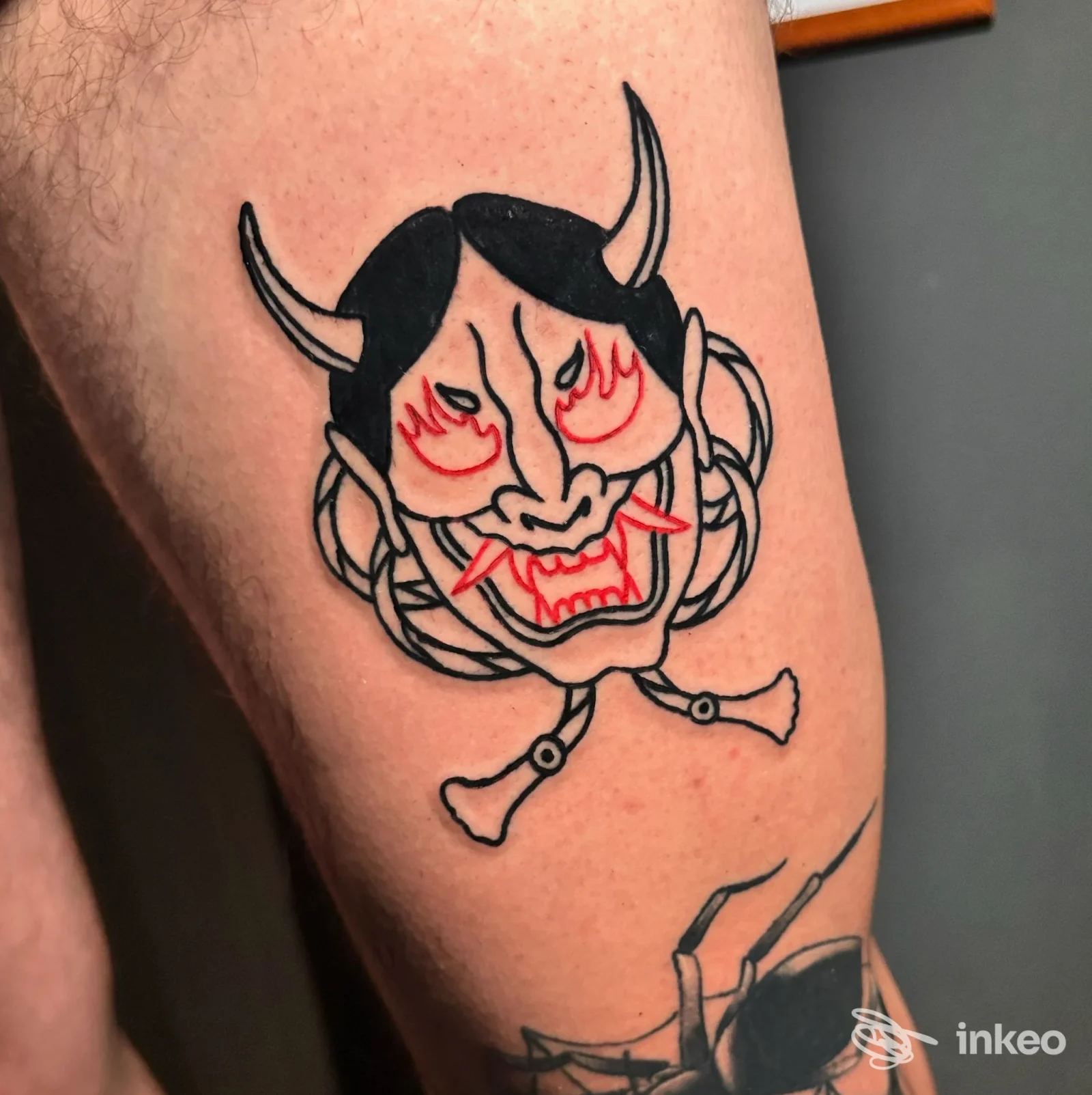 Tatuaggio a inchiostro nero e rosso raffigurante una maschera demoniaca sulla coscia, scatto dal Bunkerino Tattoo Shop, Venezia.