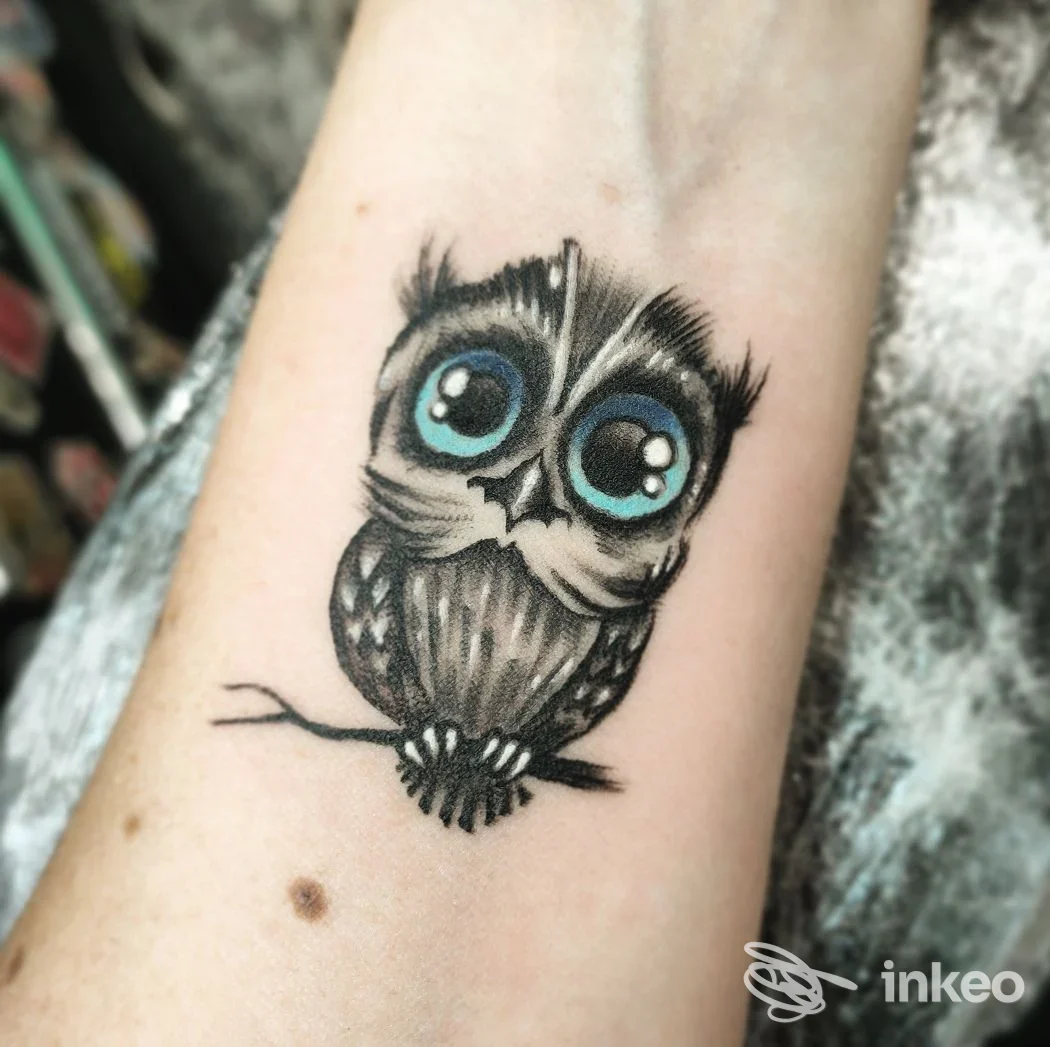 Tatuaggio di un gufo stilizzato con grandi occhi blu sull'avambraccio, realizzato da Bruce Tattoo Ink Studio Mirano.