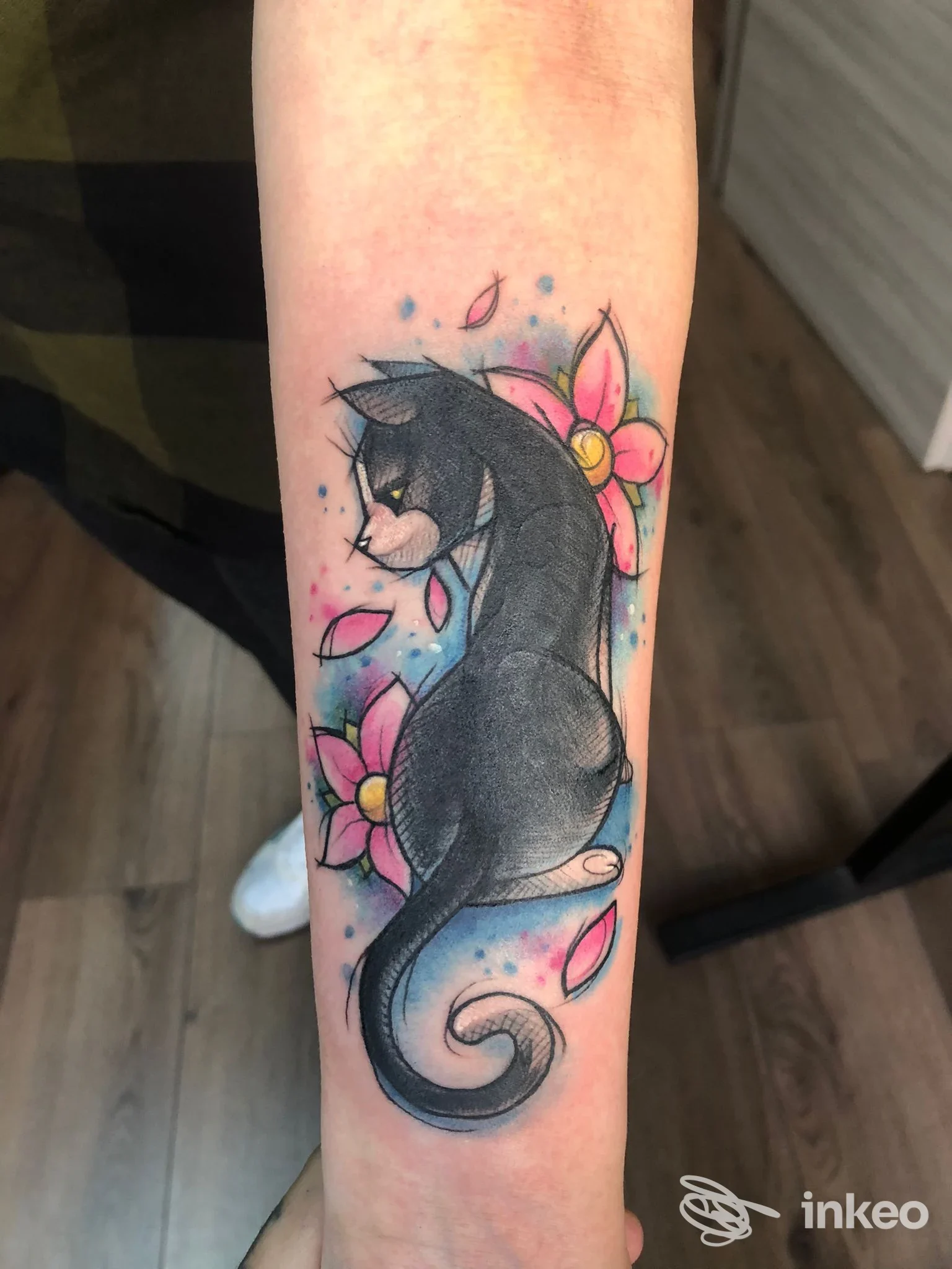 Tatuaggio su avambraccio di un gatto nero seduto tra fiori rosa e sfondo azzurro, realizzato da Scribbles Tattoo a Castano Primo.
