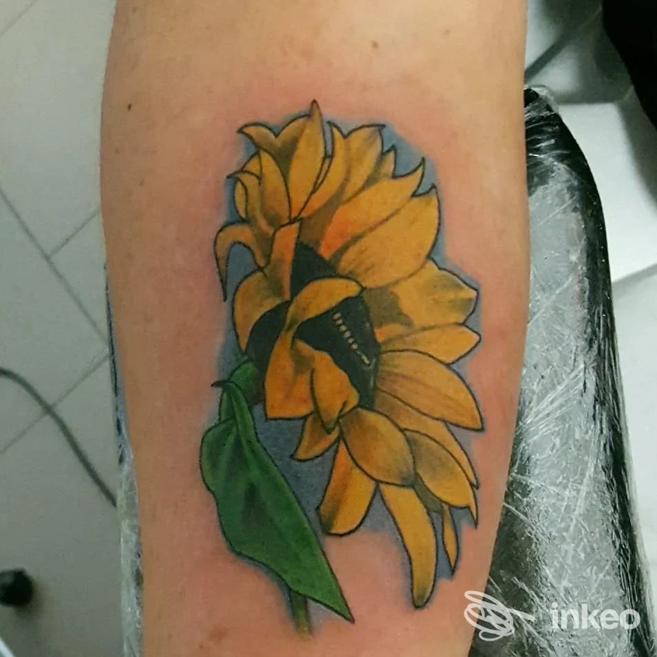 Primo piano di un girasole tatuato sull'avambraccio, colori giallo e verde su sfondo celeste, realizzato da Ink Society Tattoo a Grisignano di Zocco.