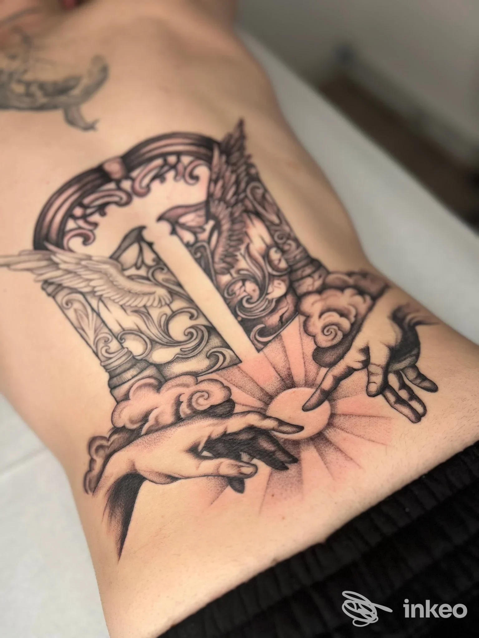 Tatuaggio ornamentale in nero/grigio sull'addome laterale con ali, elementi architettonici e mani, immagine fornita da KATRAME Tattoo Studio, Schio.