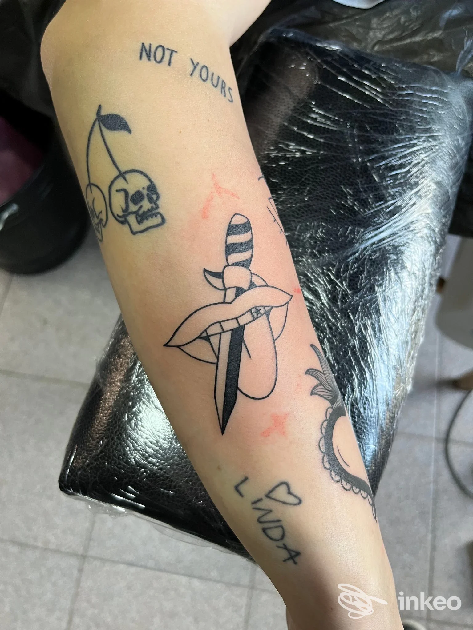 Avambraccio con più tatuaggi in nero, tra cui un pugnale attraverso una bocca; foto scattata su poggia-braccio in studio Nonsense Tattoo, Mira.
