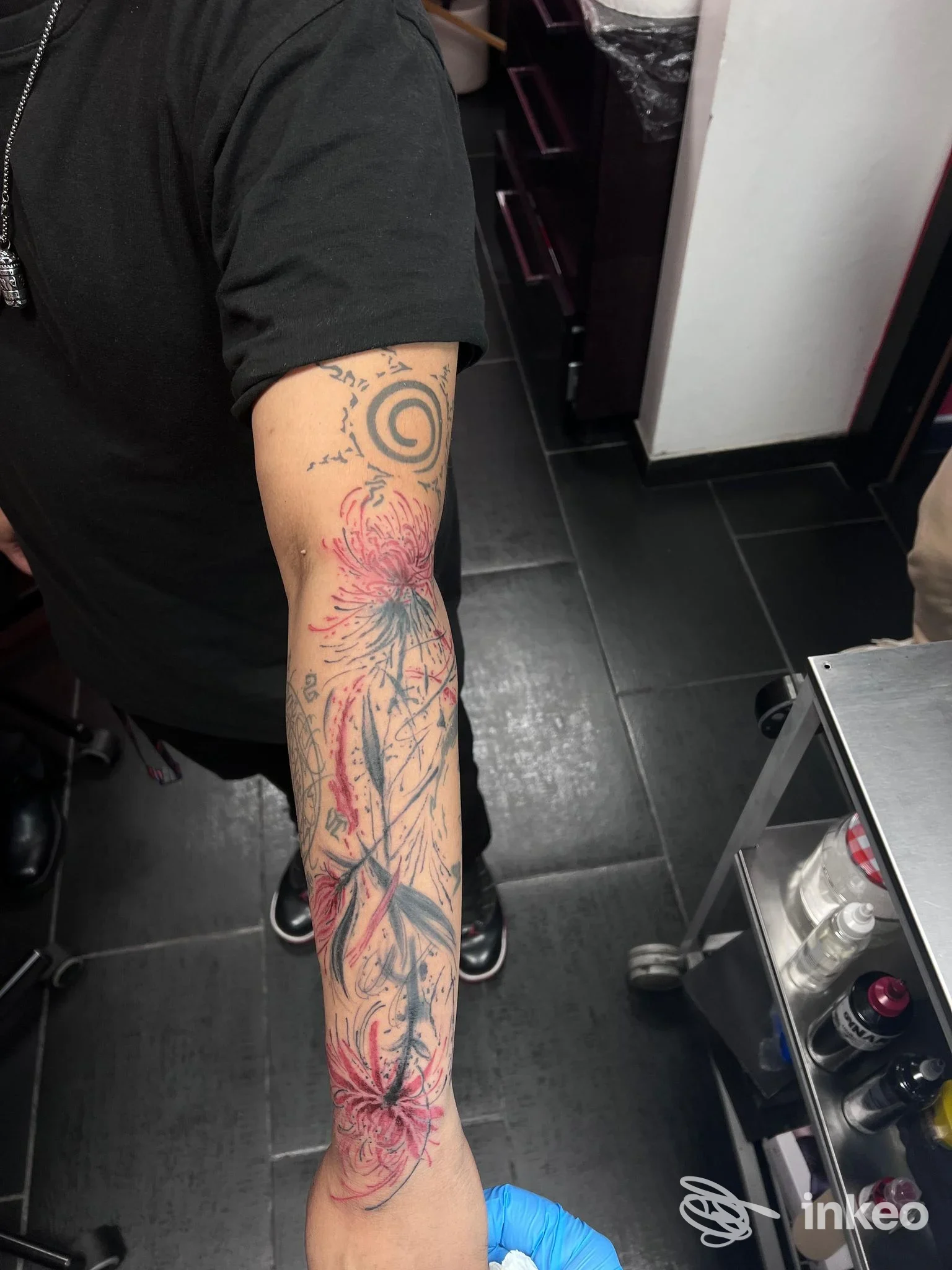 Tattoo sul braccio sinistro con motivi floreali e una spirale nera, visibile su pelle reale, sfondo di studio tatuaggi.
