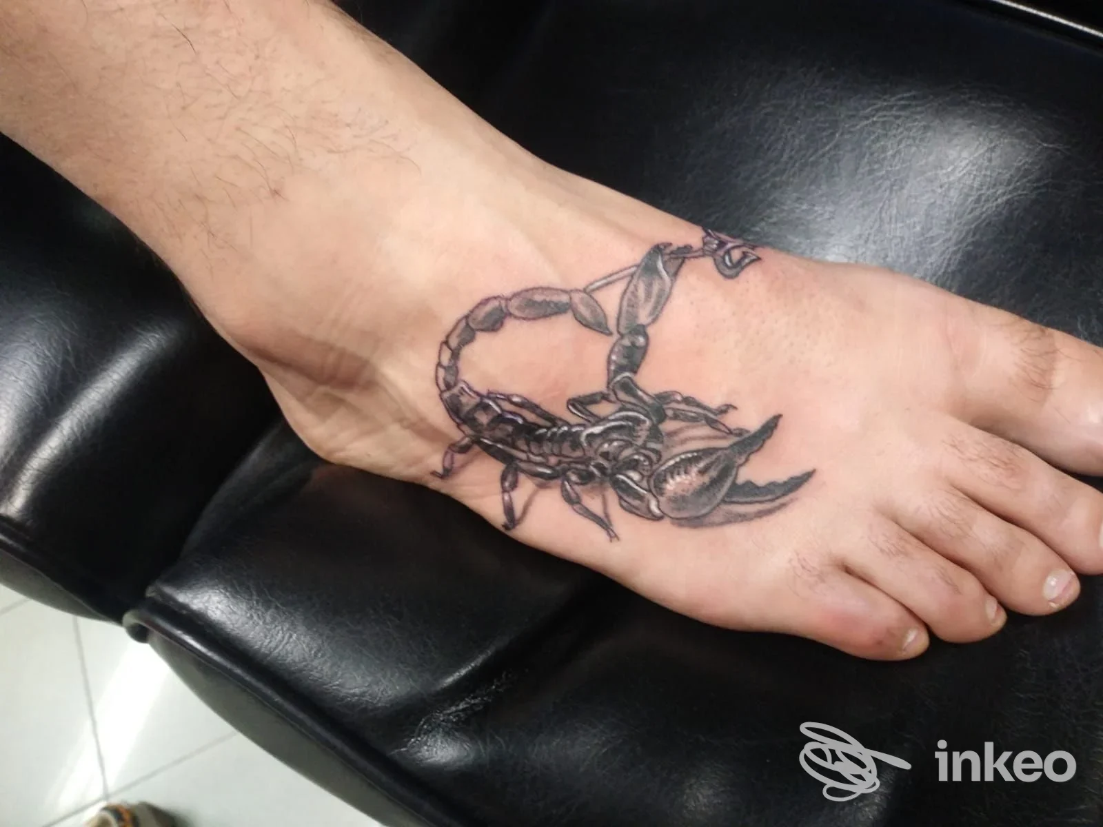 Tatuaggio realistico raffigurante uno scorpione sul dorso del piede, fotografato su sedia nera in ambiente da lavoro. Skin Painting Tattoo, Saccolongo.