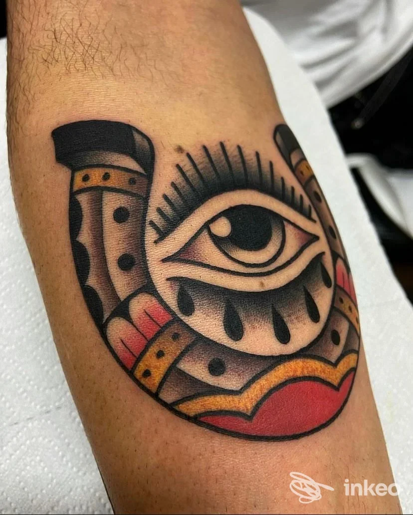 Tattoo su avambraccio raffigurante un occhio centrale incorniciato da un motivo a ferro di cavallo, colori nero, beige e rossi, contorni netti senza volto umano visibile.