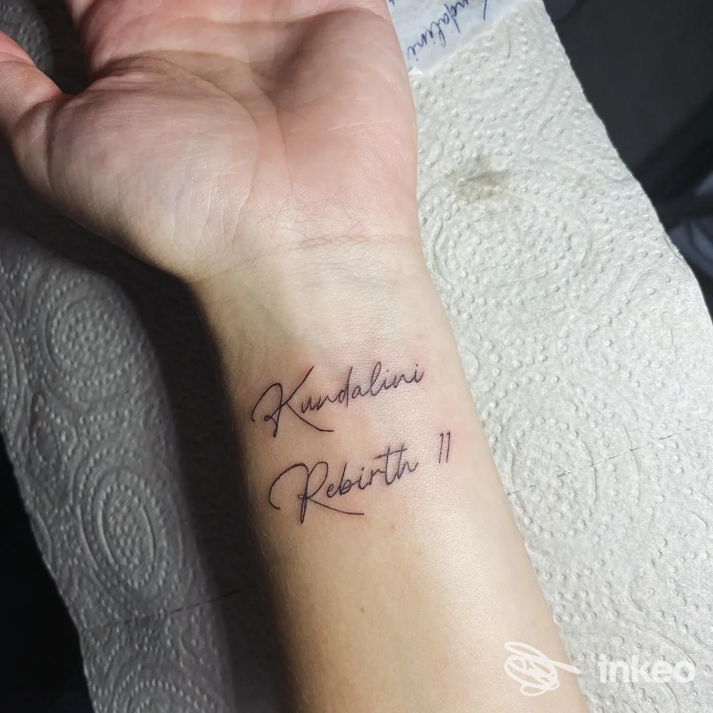 Tattoo visibile su avambraccio: scrittura in corsivo nero con parole Kundalini e Rebirth 11. Nessun volto visibile.