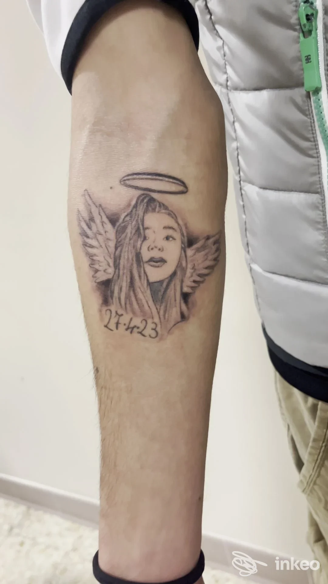Tatuaggio ritratto in nero e grigio con aureola e ali sull'avambraccio, data '2-1-23' in basso.