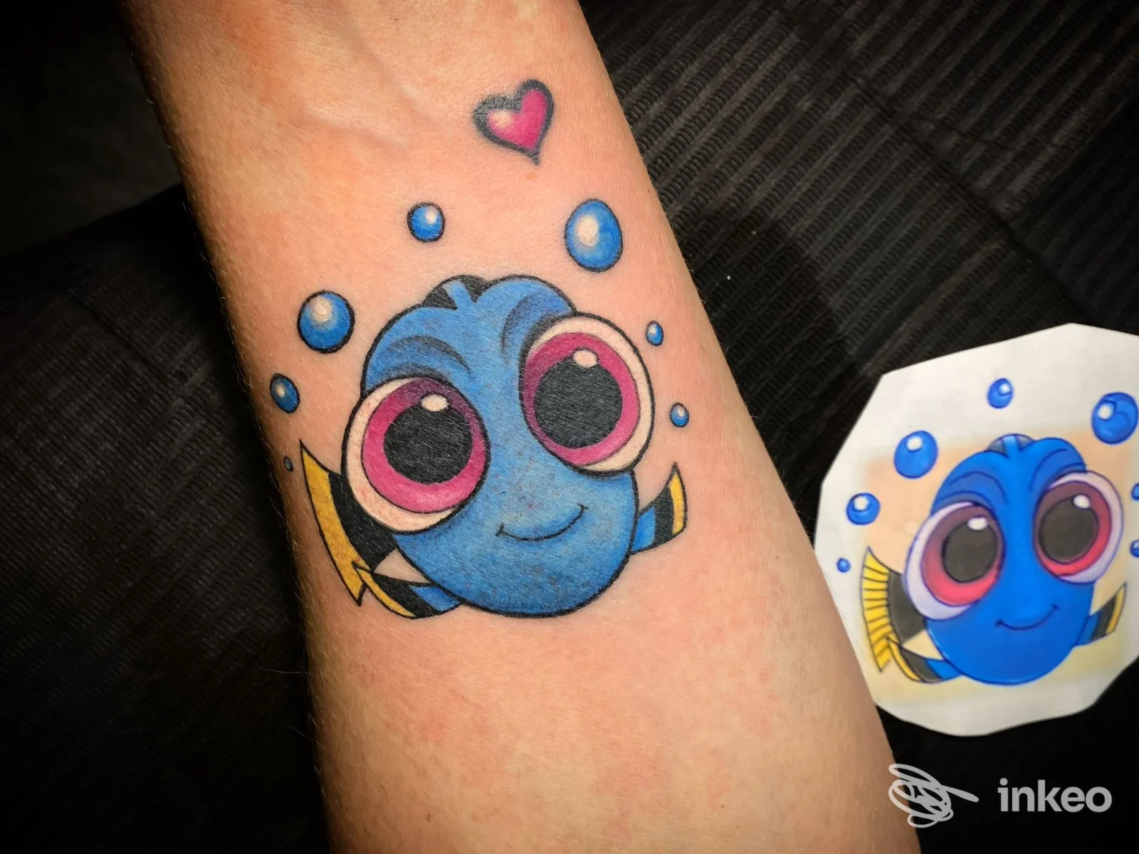 Tattoo su avambraccio raffigurante un pesce azzurro con grandi occhi rosa, circondato da bolle e un piccolo cuore rosso in alto; adesivo corrispondente visibile accanto.