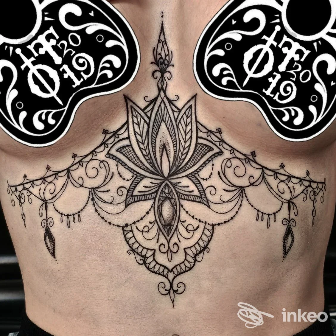 Tattoo ornamentale centrale sul torace, motivo a mandala con dettagli intricati e decorazioni pendenti lungo il petto, senza viso visibile.