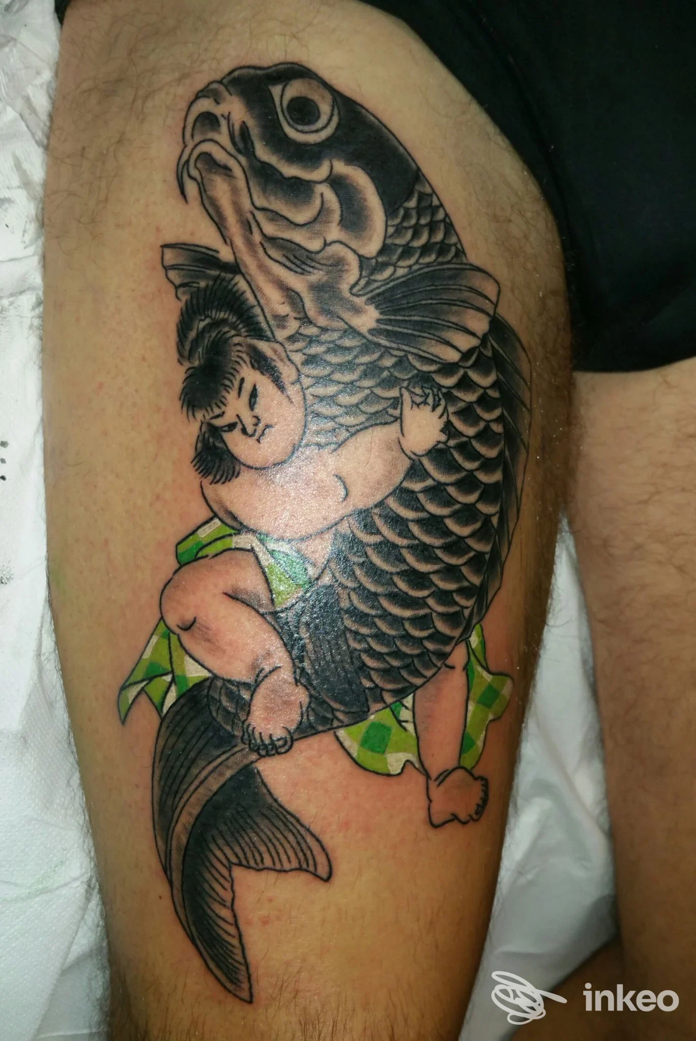 Tatuaggio su gamba raffigurante un pesce grande con squame scure e una figura femminile in abito colorato che lo avvolge, visibile solo parzialmente.