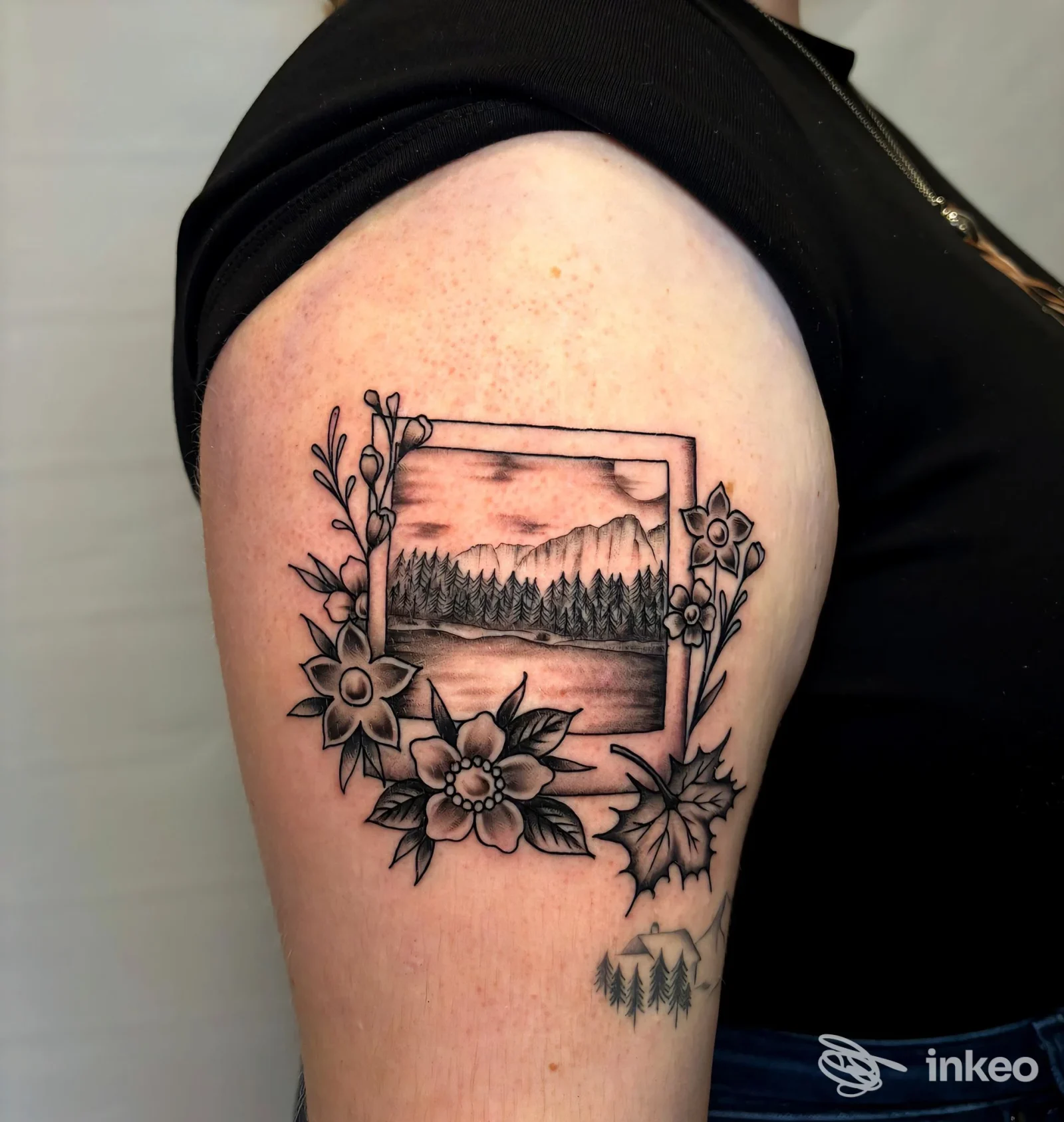 Tatuaggio in nero di paesaggio montano incorniciato con fiori sull'alto braccio, realizzato da Sacred Bones Tattoo Bolzano.