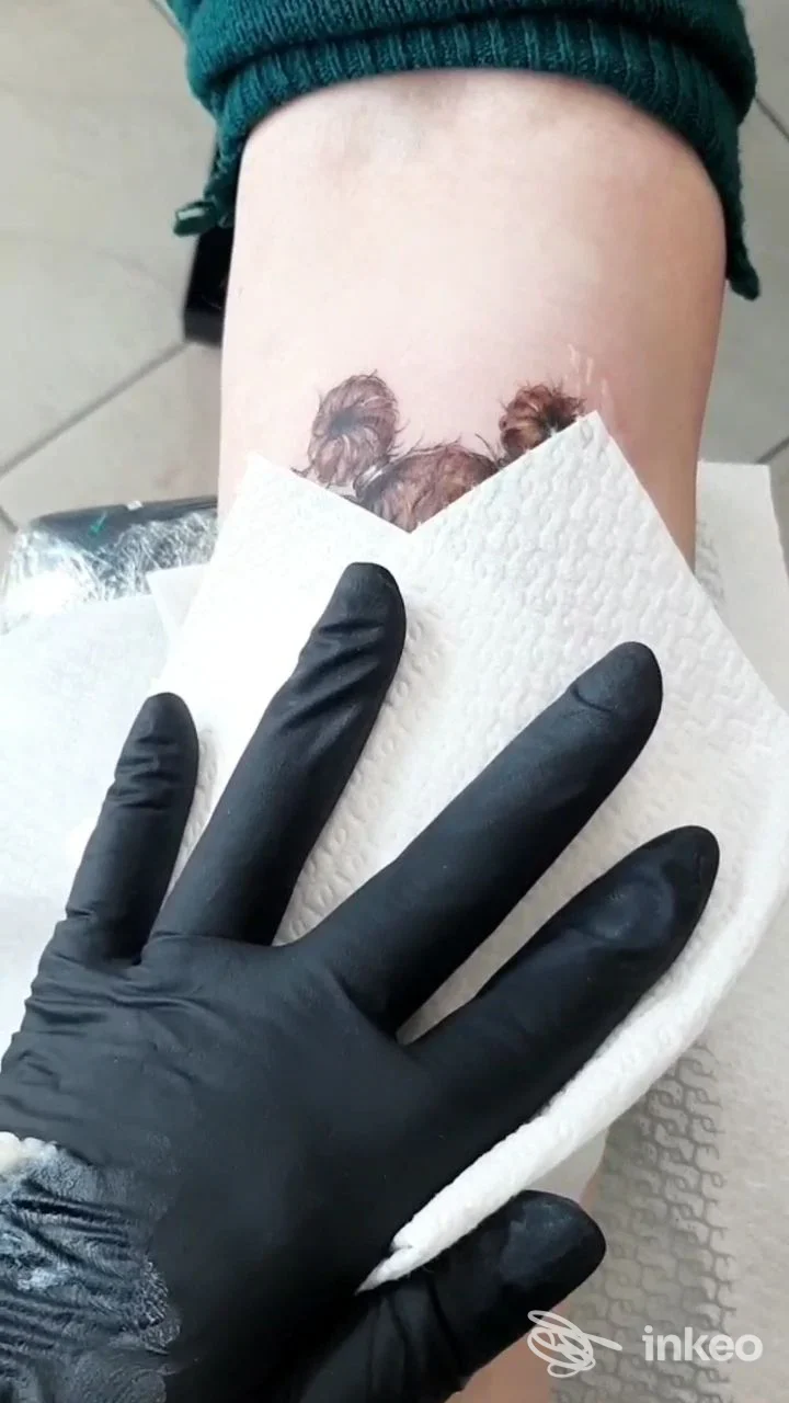 Parte del braccio con tatuaggio in fase di lavorazione, guanti neri e tessuto assorbente bianco, ambiente da studio.