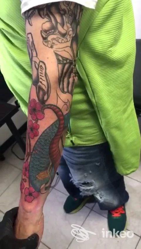 Primo piano di un avambraccio tatuato con una carpa colorata, fiori e figure; immagine fornita da Black Pyramid Tattoo, Zola Predosa.