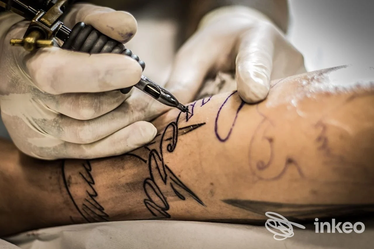 Tatuatore al lavoro su avambraccio con macchinetta: linea nera e tratti guida viola, Ink Shake Tattoo Studio, Ravenna.