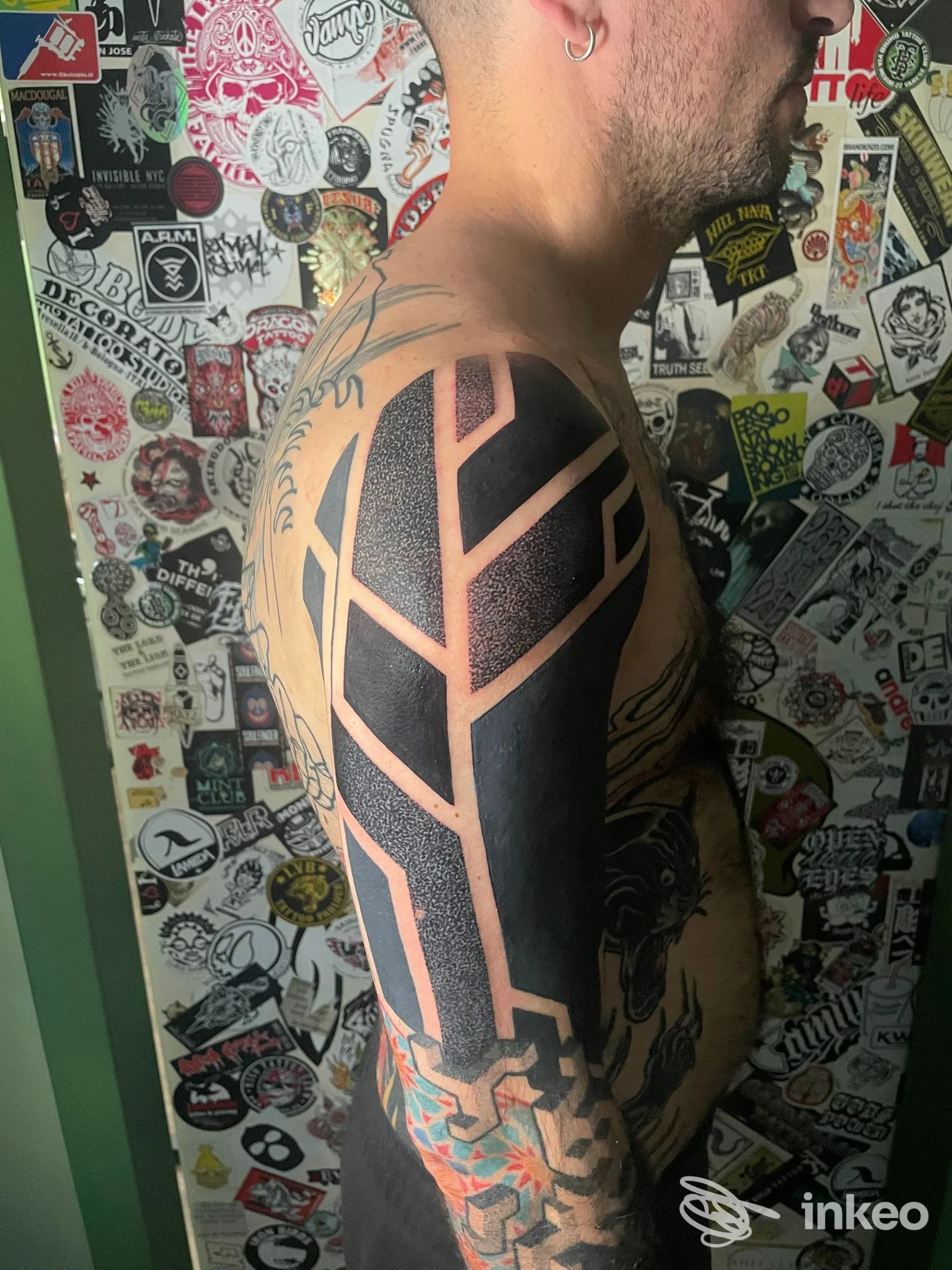 Tattoo geometrico nero su spalla e braccio di un uomo, ritratto laterale senza viso completo; sfondo ricco di adesivi.