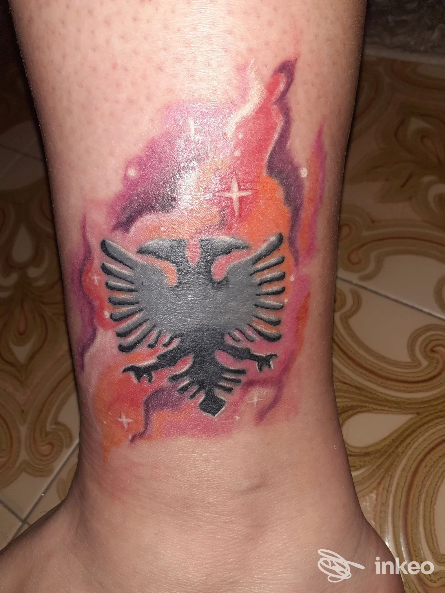 Tatuaggio aquila stilizzata su caviglia, sfondo arancio-viola. Glory Bound Ink Tattoo Shop & Piercing, San Lazzaro di Savena.