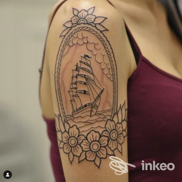Tattoo sul braccio raffigurante una vela in una cornice ovale di corde con fiori ai margini inferiori, eseguito a inchiostro nero sulla pelle.