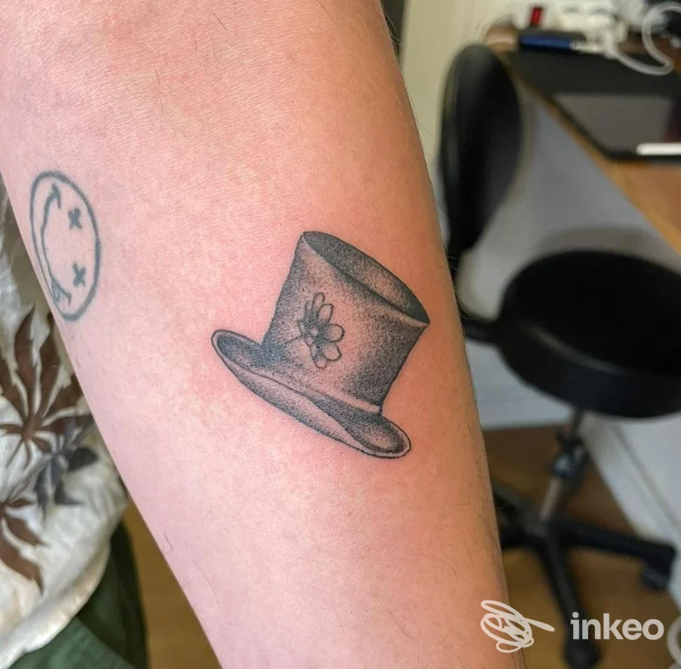 Avambraccio con due tatuaggi, cappello a cilindro nero/grigio e piccolo volto stilizzato, Out of Line tattoo studio Bologna