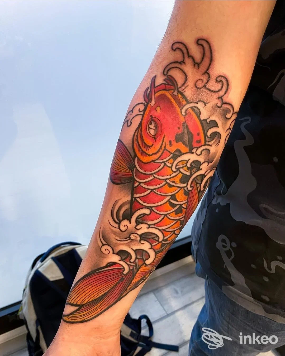 Avambraccio visibile mostra un tatuaggio di carpa koi con onde, contorni scuri e colori vivaci; la pelle è ben definita e la grafica copre una porzione significativa dell’avambraccio.