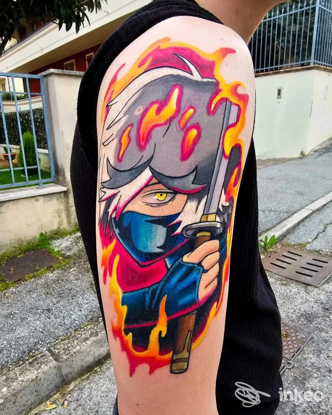 Tatuaggio a colori sul braccio superiore raffigurante un personaggio mascherato con katana — immagine fornita da Ame Noire Tattoo Shop, Riccione.