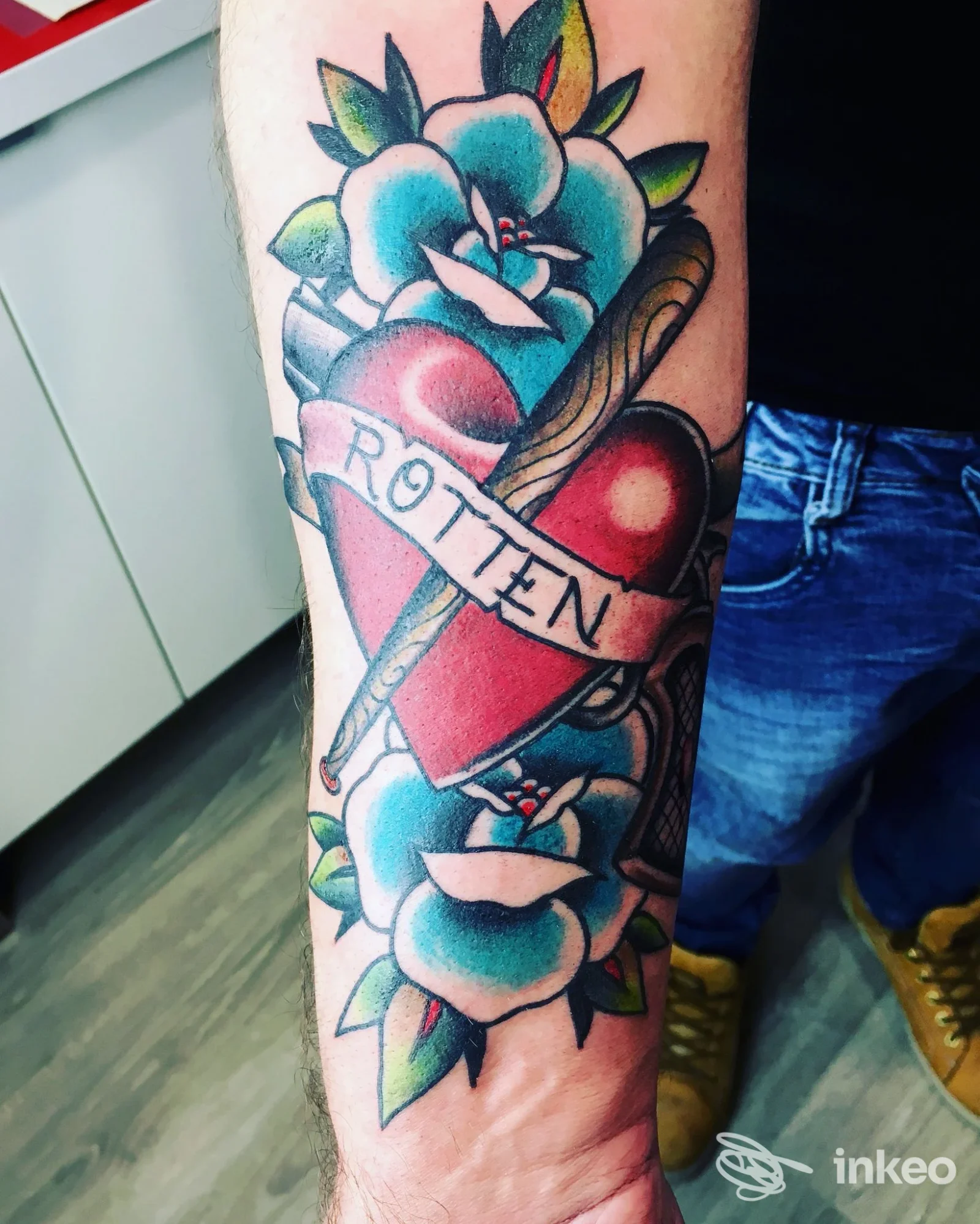 Tattoo a colori su avambraccio: cuore rosso con spada e una fascia con la scritta ROTTEN, contorni e fiori azzurri intorno.