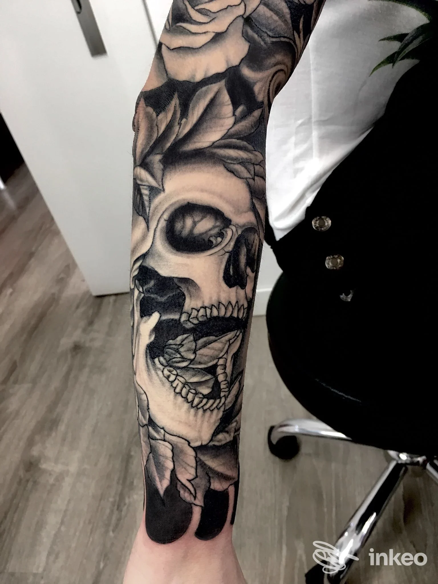 Braccio sinistro con tatuaggio realistico a tema teschio e rose, realizzato su pelle, in bianco e nero, sullo sfondo studio.
