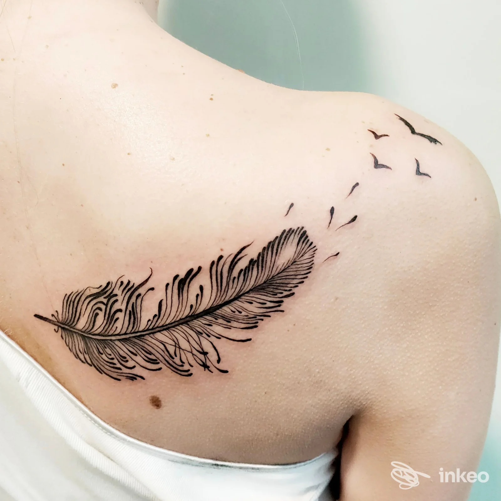 Tatuaggio di una piuma stilizzata con piccoli uccellini che volano, linework nero sulla parte superiore della schiena; immagine per SilviaRossi artgallery a Bibbiena.