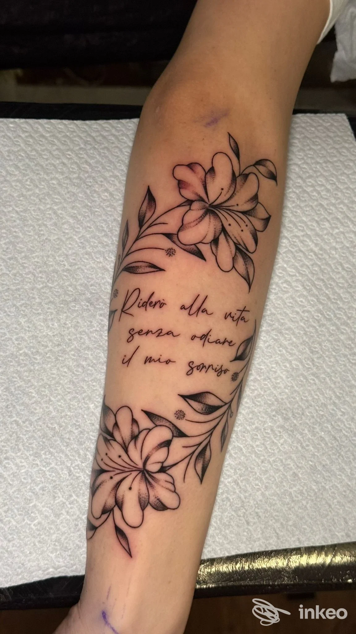 Tattoo floreale su avambraccio con due grandi fiori e scritta corsiva al centro, senza elementi di interfaccia o testo promozionale visibile.