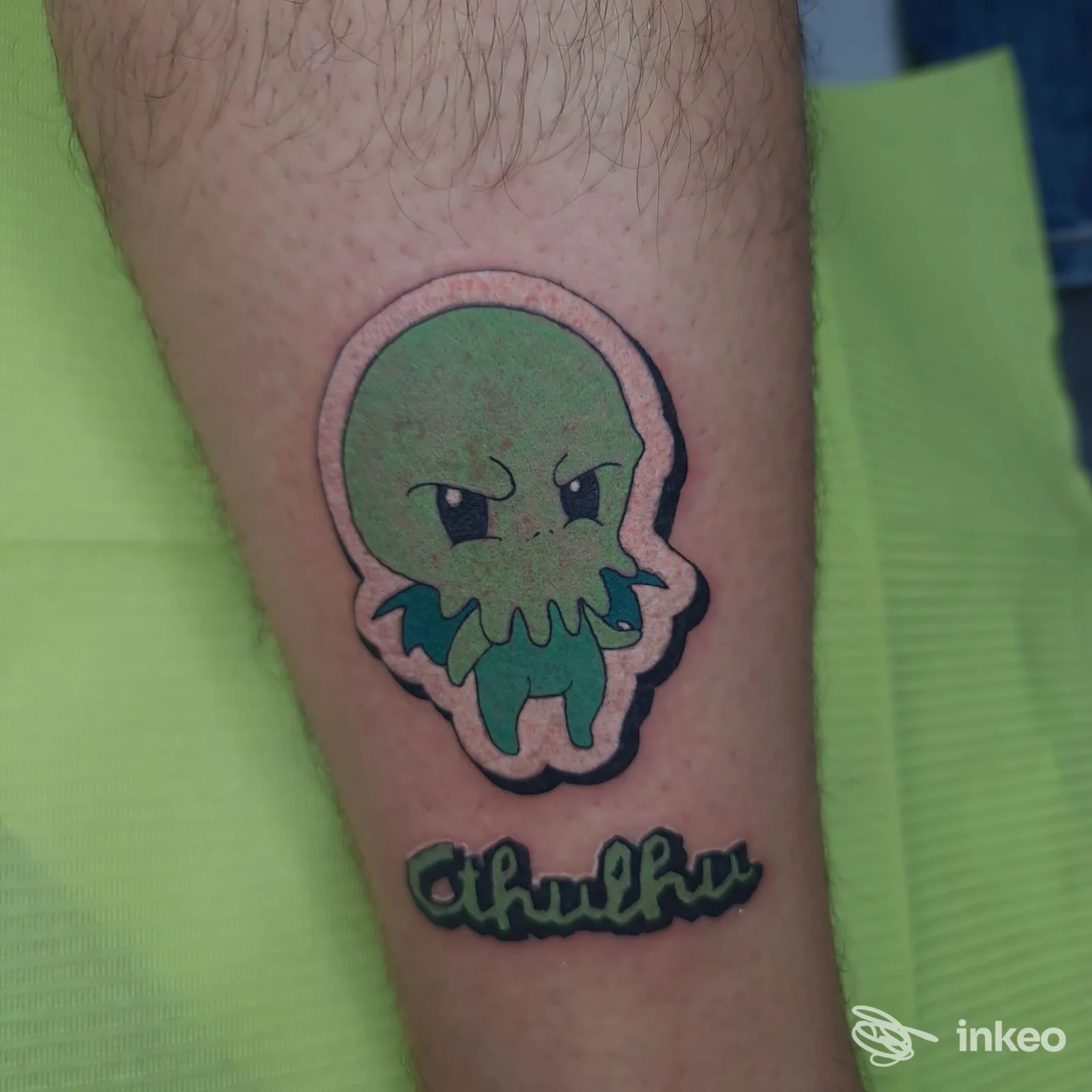 Tatuaggio colorato su avambraccio: mascotte/cartoon verde con contorno spesso e scritta "Cthulhu". Tuscan Tattoo Studio, Prato.