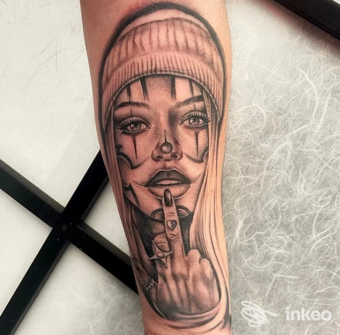 Tatuaggio in bianco e nero su avambraccio: volto femminile con berretto e mano reale con dito medio, HOMEBODY Tattoo Lab, Bologna.