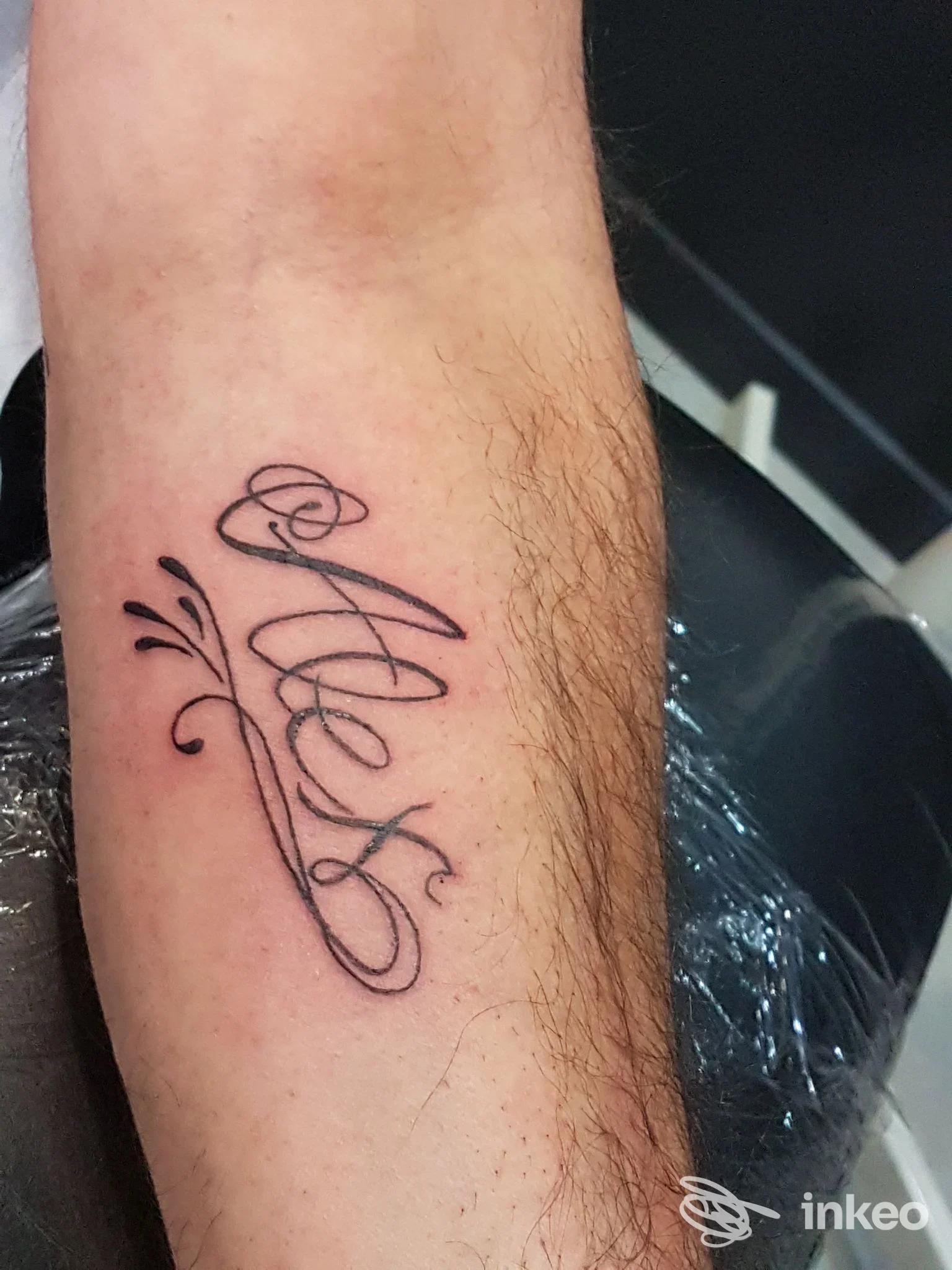 Tatuaggio in stile calligrafico su avambraccio sinistro, linee sottili nere e decorazioni laterali; opera corporea reale visibile su pelle.