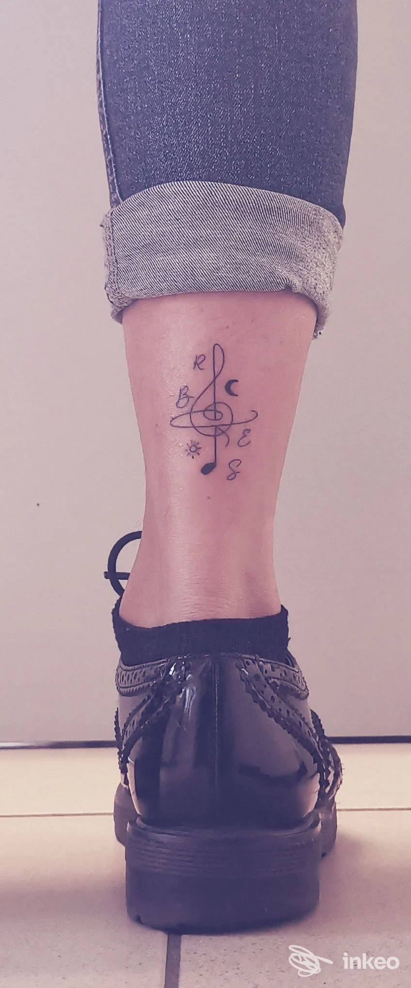 Ginocchio e caviglia destra con pantalone arrotolato, mostra un tatuaggio astratto sull’osso della caviglia, di linea nera/blu, in primo piano accanto a una scarpa nera lucida.