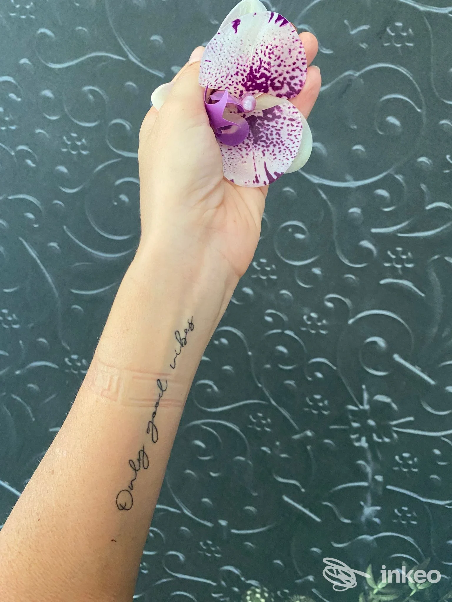 Tattoo sull’avambraccio eseguito presso lo studio Paola Bazzani: testo nero lungo la pelle, mano che tiene un fiore orchid violaceo su sfondo decorativo.