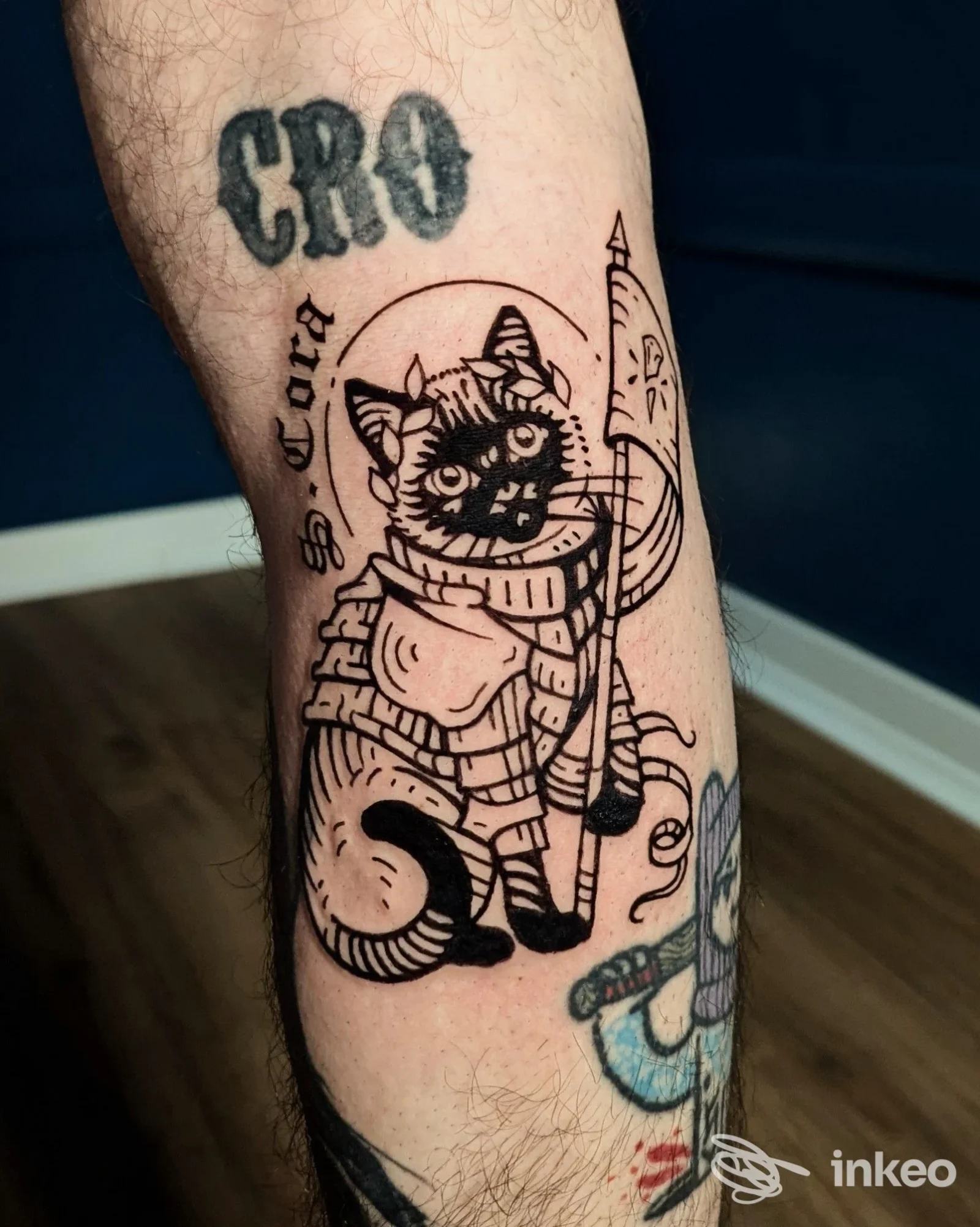 Foto di un tatuaggio a Torino presso Sacrosanto Tattoo: gatto in armatura in nero sul polpaccio, con altri tatuaggi circostanti.
