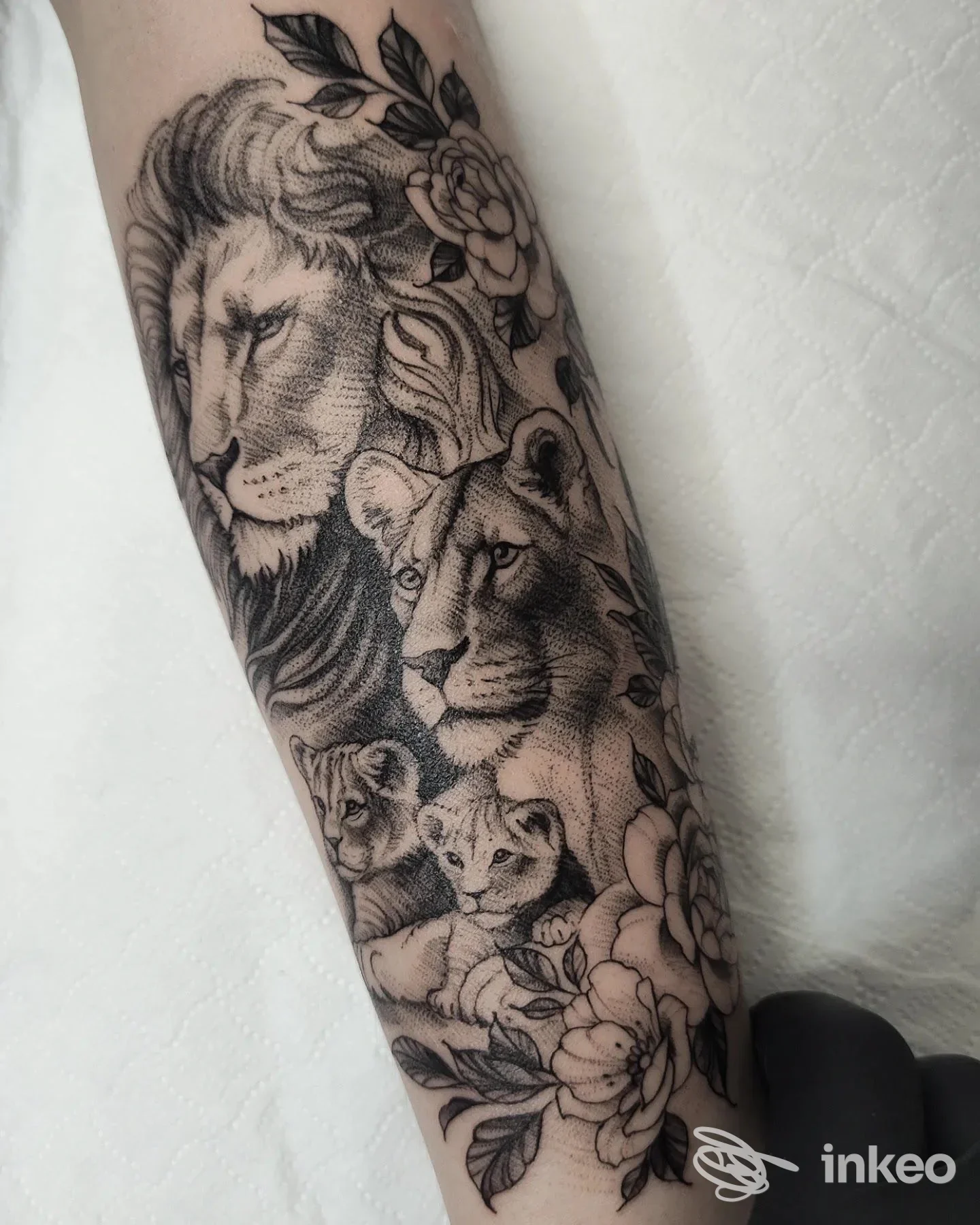 Tattoo sull'avambraccio raffigurante un leone realistico in bianco e nero con altri leoni cuccioli e fiori, braccio completamente visibile.