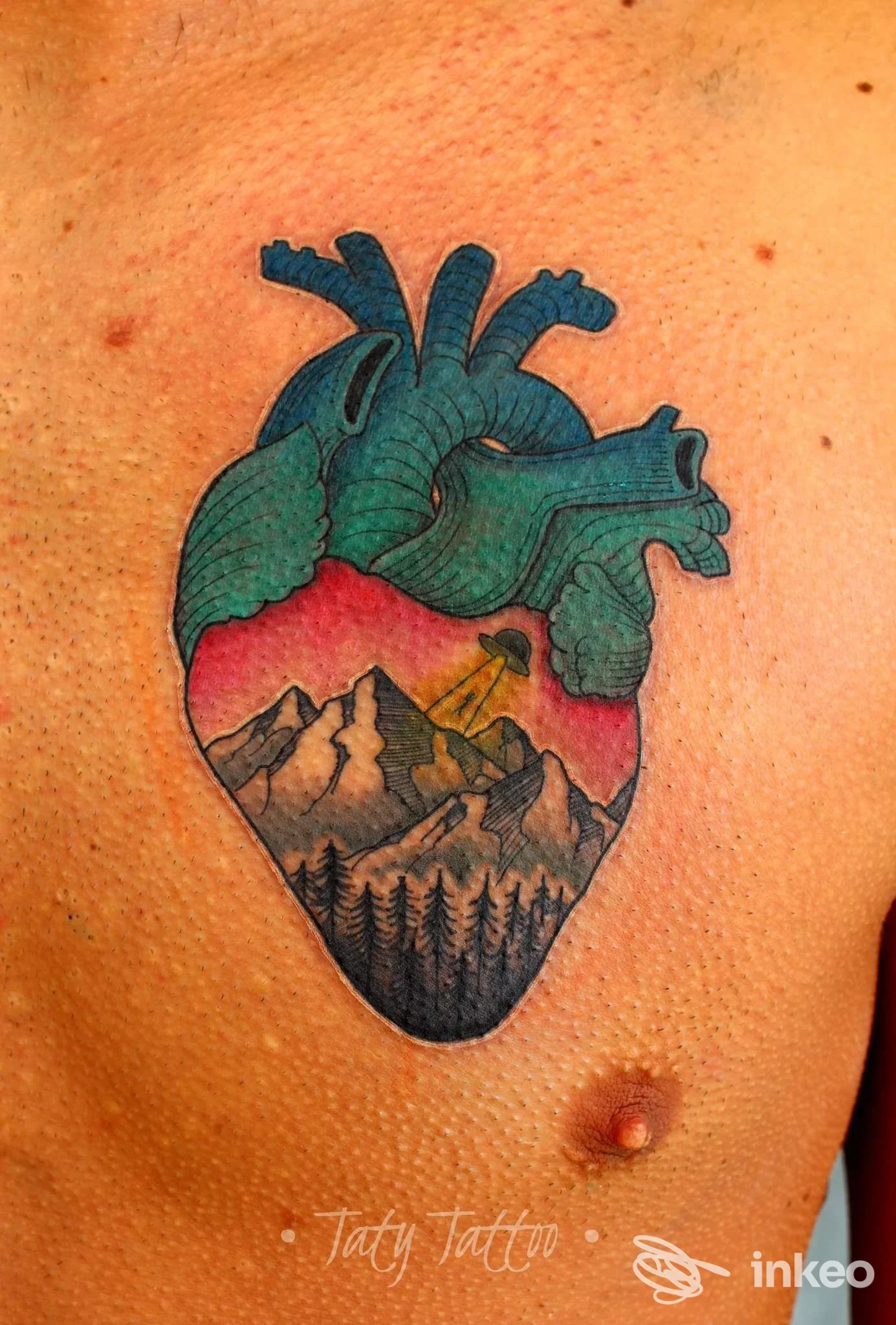 Tattoo a cuore sul petto sinistro, raffigurante paesaggio montuoso con foresta e cielo rosato, colori verdi/blu e contorni neri.